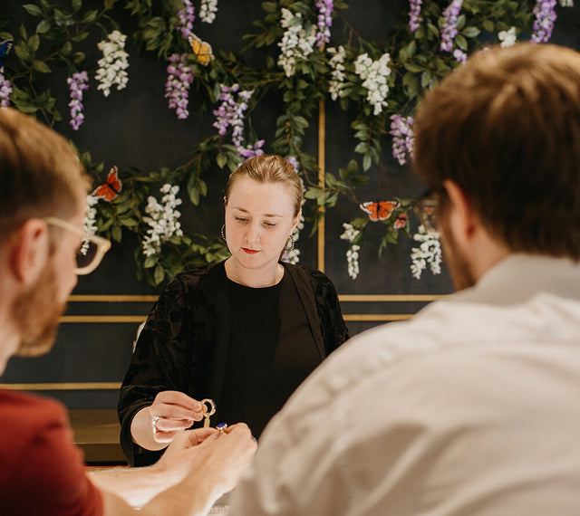 a sales women showing two male customers a ring. There is a floral display behind her.